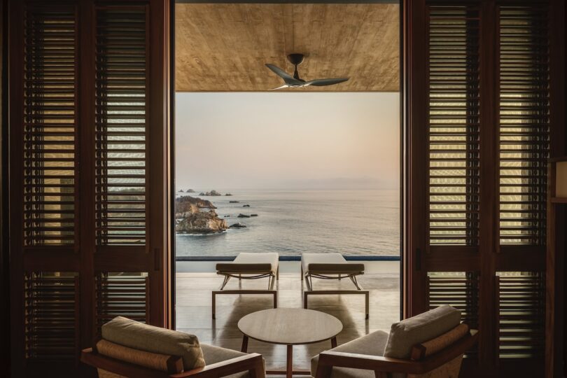 View from a modern room through open wooden doors to a balcony with two lounge chairs overlooking a calm ocean and rocky coastline.