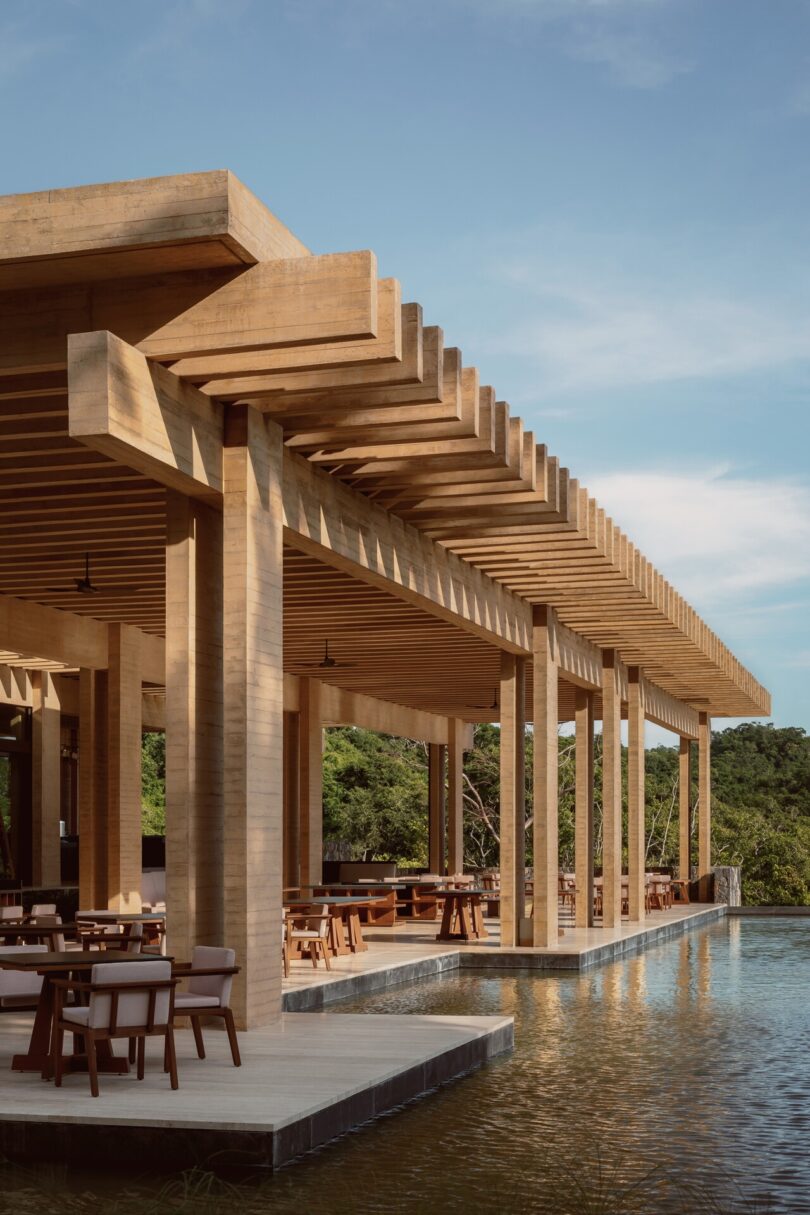 A wooden pavilion with a pergola roof extends over a reflective water surface, surrounded by trees, under a clear blue sky.