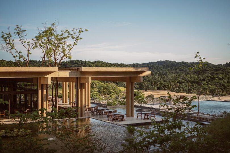 Modern open-air building with wooden pillars beside a reflecting pool, surrounded by lush greenery and overlooking a hilly landscape. Chairs and tables are placed beneath the structure.