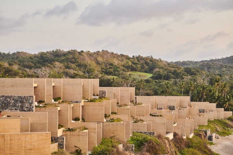 A series of modern, beige concrete buildings with flat roofs situated on a hillside, surrounded by lush greenery and trees under a cloudy sky.