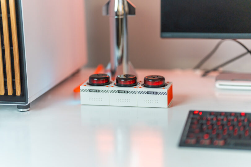 A desktop setup featuring a monitor, keyboard, and three cylindrical devices on a white desk.