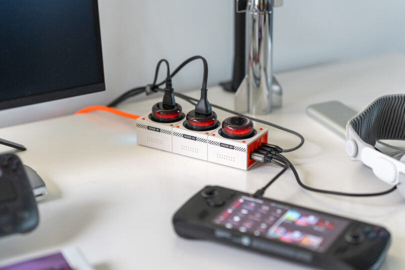 A desk with a power strip featuring multiple orange and black circular outlets, connected devices, a monitor, headphones, and a handheld gaming device.