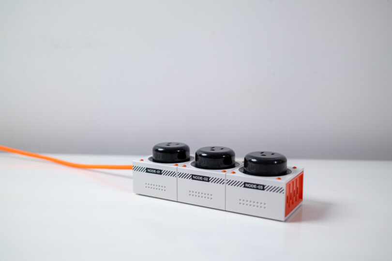 A power strip with three black sockets and an orange cable is placed on a white surface against a plain background.