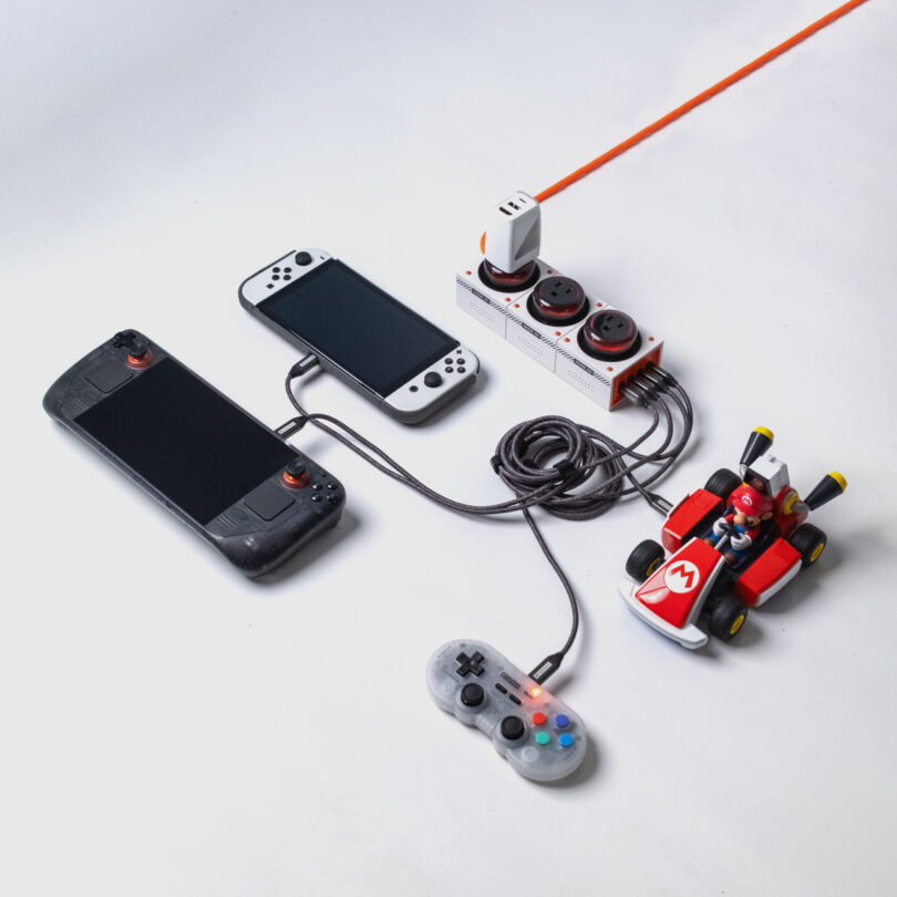 Two handheld gaming consoles, a wired controller, a power strip with plugged-in adapters, and a toy race car with a driver on a white surface.