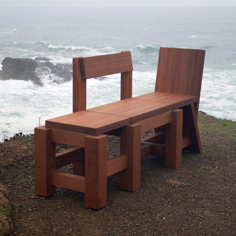 Wooden bench with tall backrest on one side, placed on a dirt path overlooking ocean waves and rocky shore. Overcast sky in the background