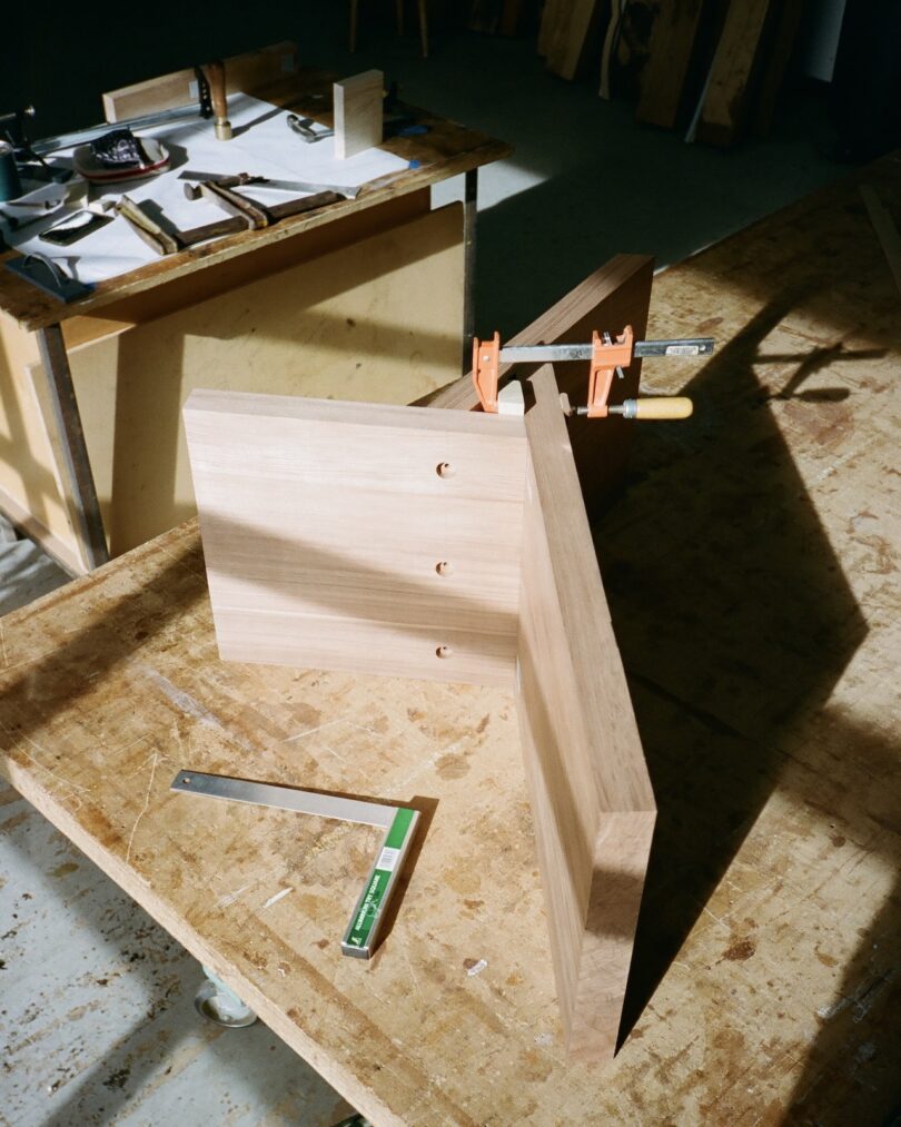 Woodworking bench with clamps holding wooden pieces in place. Tools and a ruler are on the surface. Sunlight casts shadows across the workbench