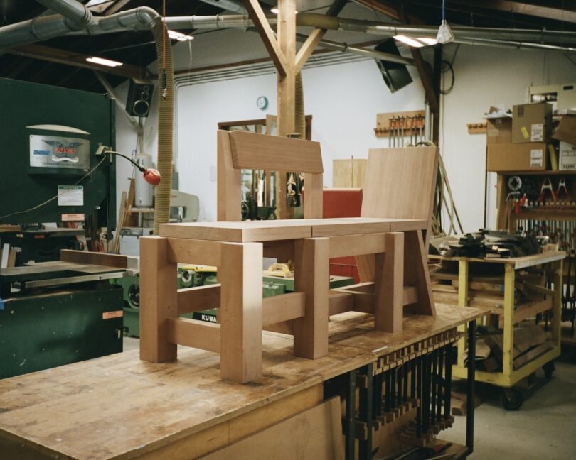 Wooden chairs under construction sit on a workbench in a cluttered workshop with tools and equipment visible in the background