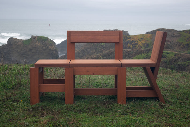 Wooden bench with an angular design sits on a grassy area by the coast, with rocky cliffs and ocean waves in the background.