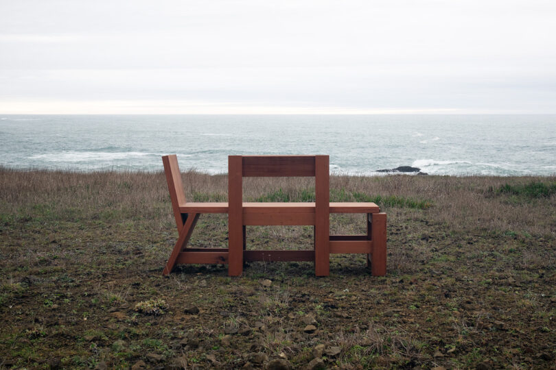 A wooden bench with back-to-back seats stands on a grassy area, overlooking the ocean under a cloudy sky