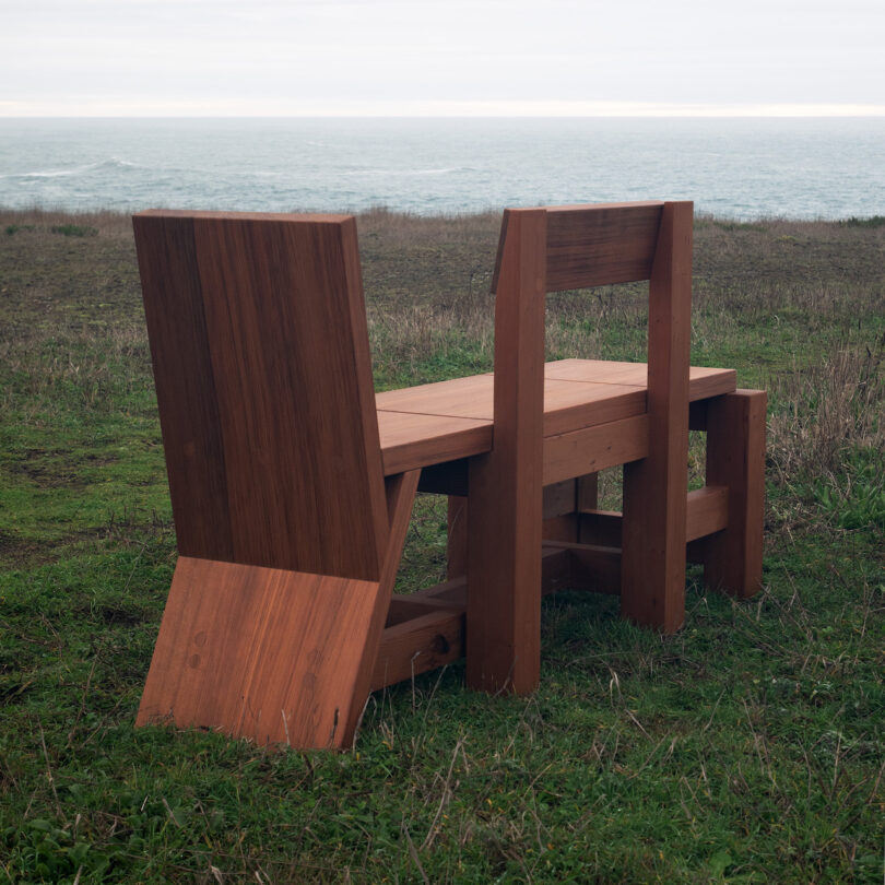 Two modern wooden chairs with tall backs, situated on a grass field overlooking the sea