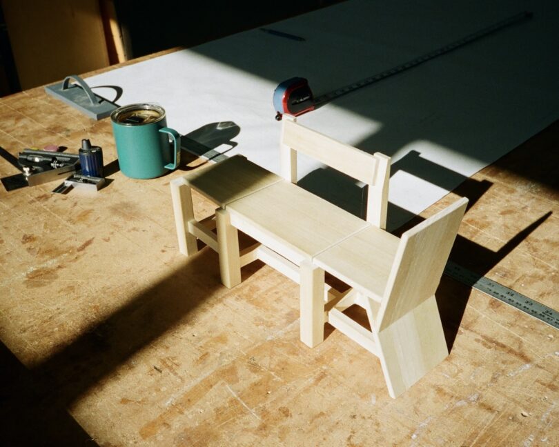 A small, light wood chair model on a wooden workbench with a measuring tape, tools, a cup, and a blueprint in the background