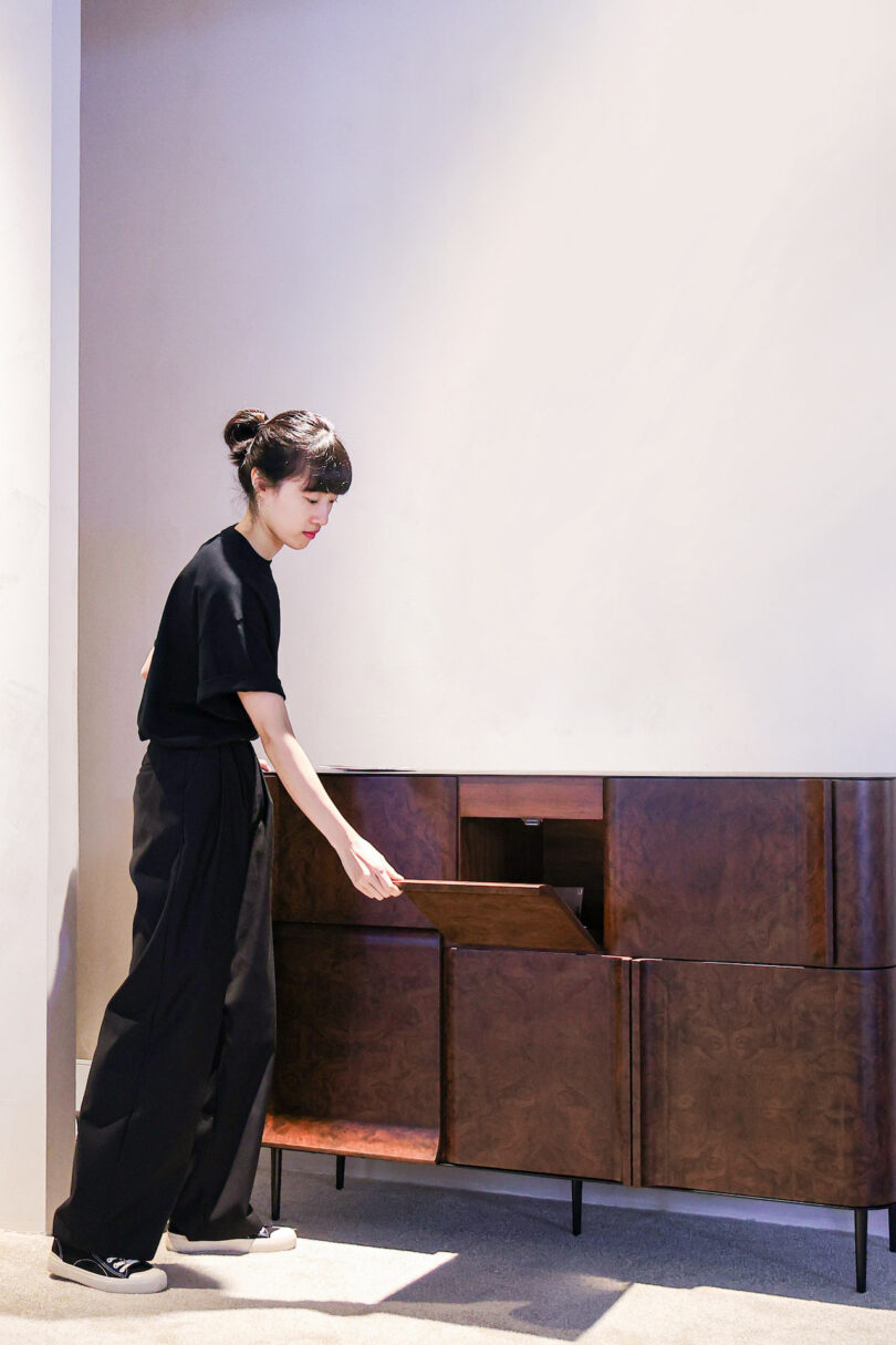 Person in black outfit opens a wooden cabinet door in a bright room