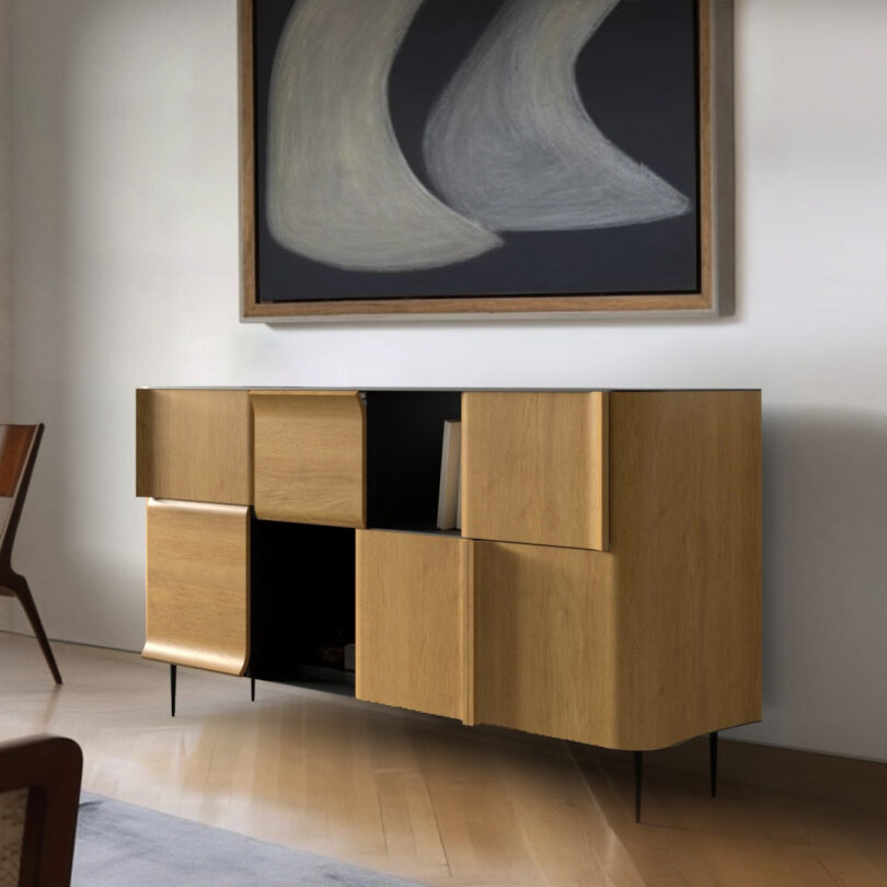 A modern wooden sideboard with open, geometric cabinets stands on a wooden floor. Above it hangs a large abstract painting in black and gray