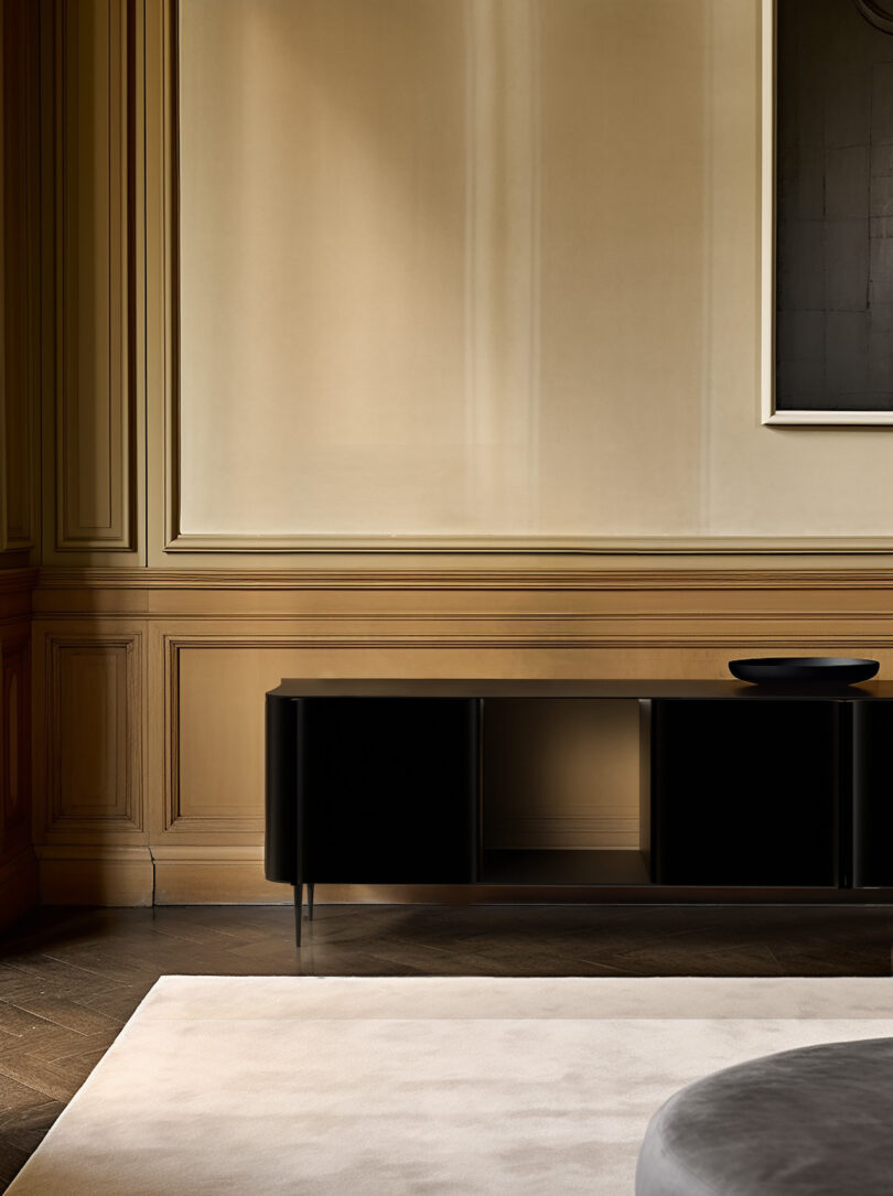 A minimalist interior with beige walls, a black console table, and a black-framed artwork. The floor has dark wood, and a light-colored rug is partially visible in the foreground