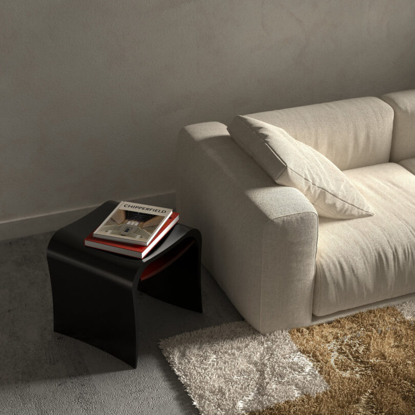 A white couch with a pillow is next to a black side table holding two stacked books in a living room. A textured rug is partially visible on the floor