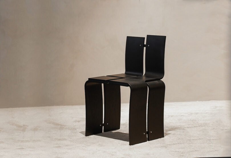 A modern chair with a unique design featuring two connected black curved metal seats, placed on a light carpeted floor against a neutral wall