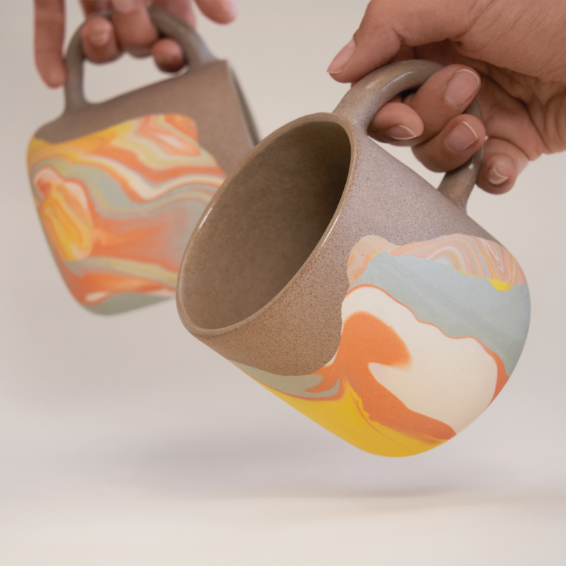 Two hands holding ceramic mugs with abstract orange, yellow, and white patterns against a neutral background