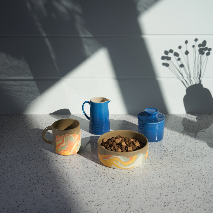 Ceramic mug, bowl with kibble, blue pitcher, and blue jar on a speckled countertop with a shadow of a vase holding dried flowers in the background