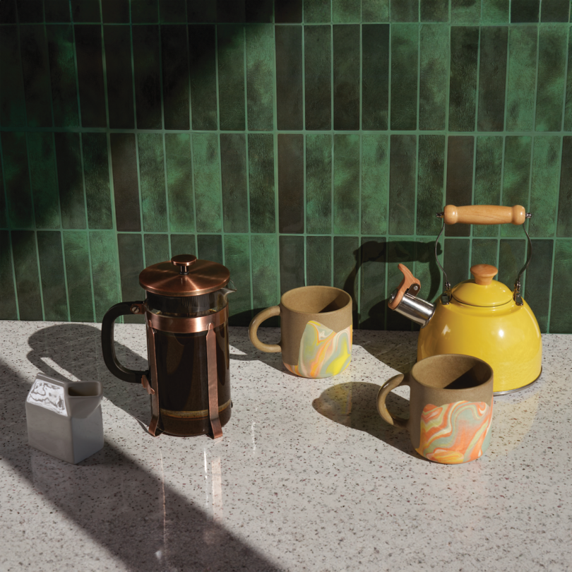 A coffee press, two mugs, a yellow teapot, and a small carton sit on a speckled countertop with a green tiled wall in the background