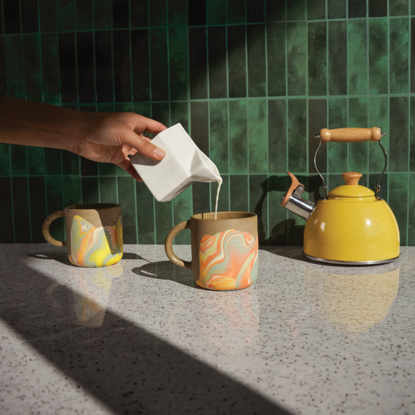 A hand pours milk from a small pitcher into one of two colorful mugs on a speckled countertop. A yellow kettle sits nearby against a green tiled wall