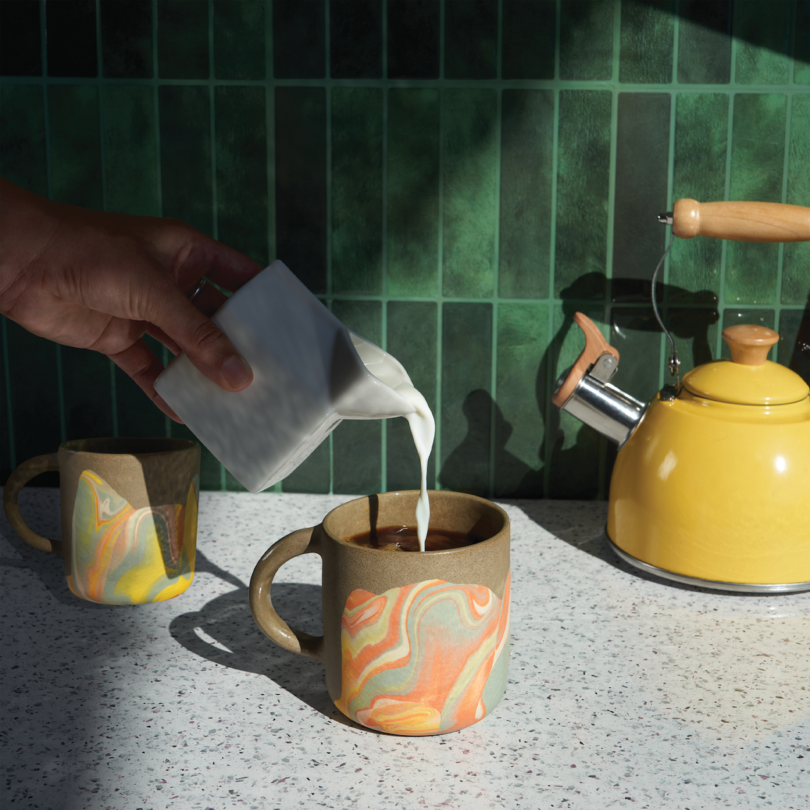 Pouring milk into a marbled mug of coffee on a speckled counter. Another mug and a yellow kettle are in the background. Sunlight casts shadows on the green tiled wall