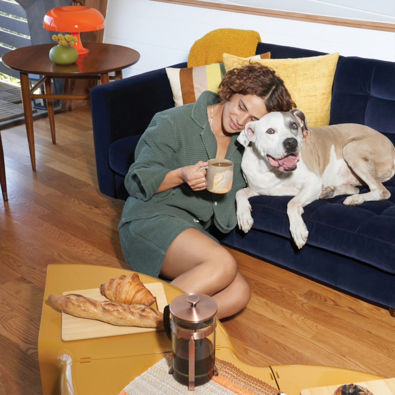 A woman sits on a blue couch with a dog, holding a mug. A coffee table in front has croissants, a baguette, and a French press. A window and orange lamp are in the background