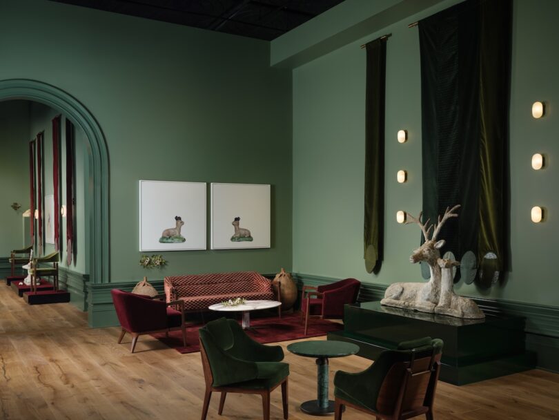 A green room with velvet chairs and a couch, deer sculpture, two wall paintings, tall curtains, and wooden flooring.