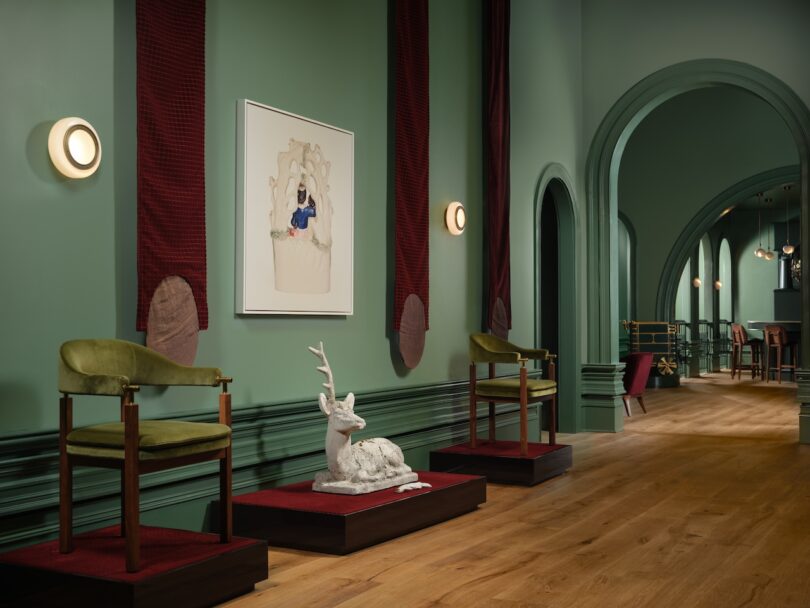 Green-walled room with wooden flooring, featuring a sculpture of a deer, two green chairs on platforms, and a framed artwork on the wall.