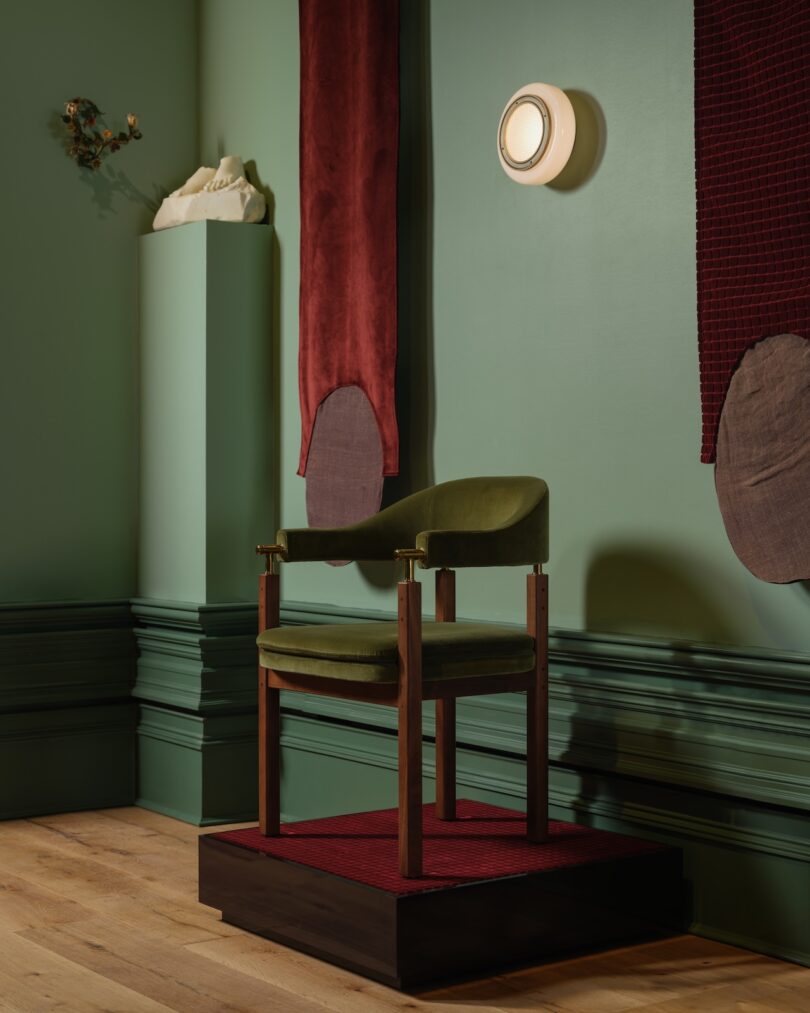 A green chair with wooden arms is placed on a red platform. It is surrounded by green walls, red fabric panels, and a white wall-mounted light. A small sculpture is displayed nearby.