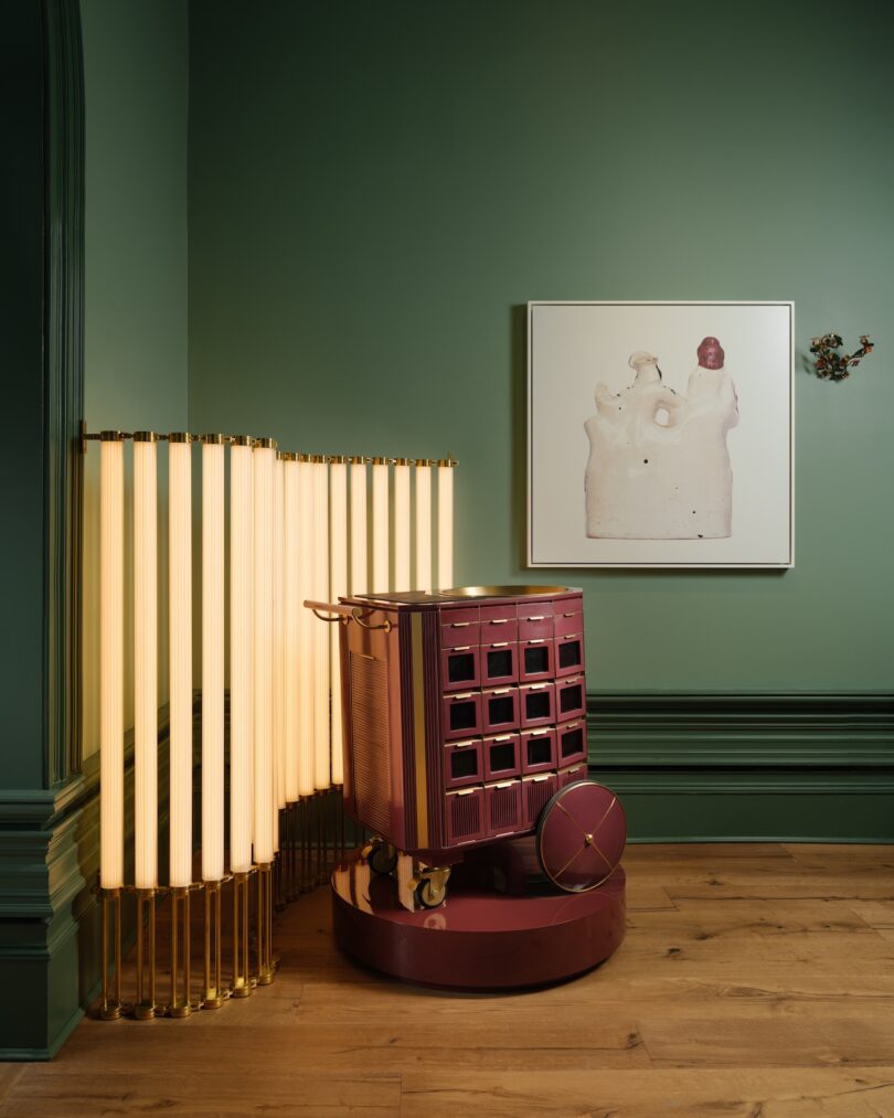 A burgundy rolling cabinet next to a tall, vertical light fixture, set against a green wall with a framed abstract art piece and a small wall decoration.