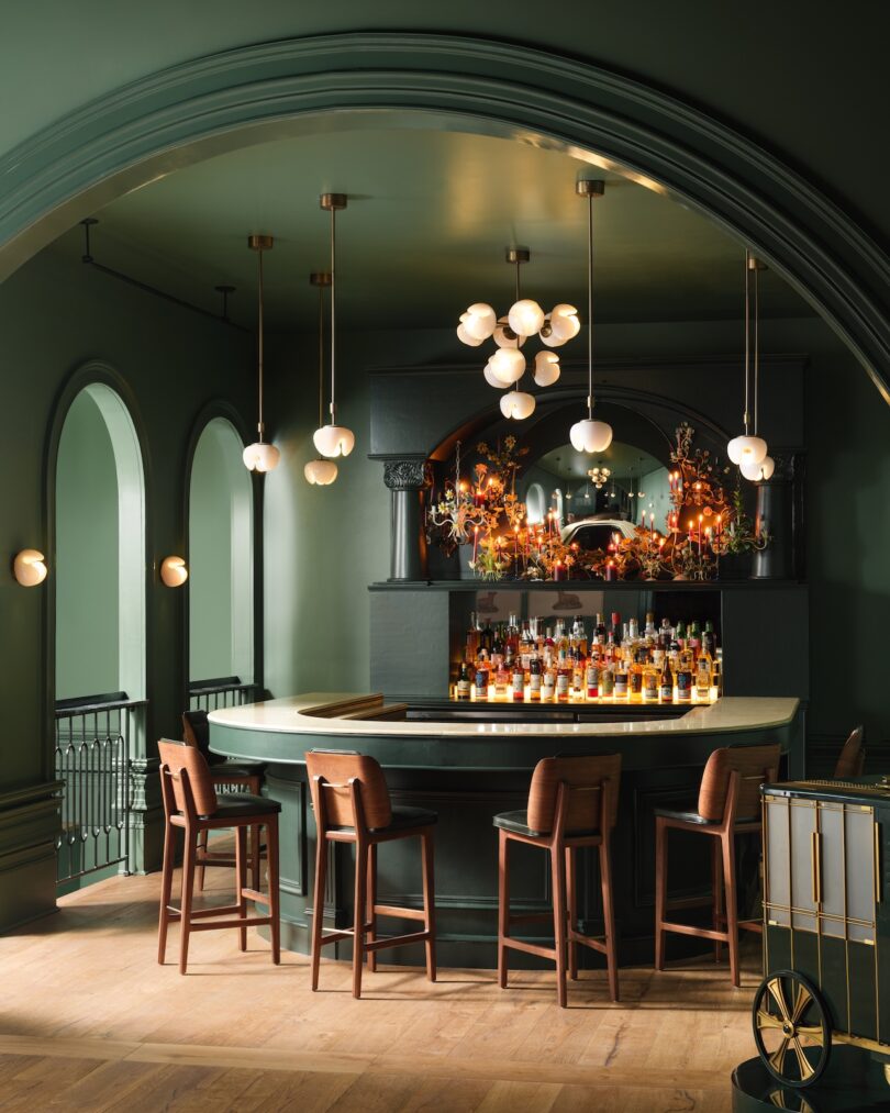 A stylish bar features a curved counter with stools, pendant lighting, and various bottles on shelves. Arched doorways and dark green walls enhance the elegant ambiance.