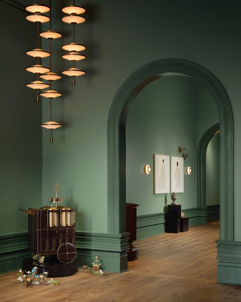 Green-painted room with arched doorway, modern chandelier, wooden floor, and decorative items on the side. Two framed artworks and small sculptures are visible in the hallway.