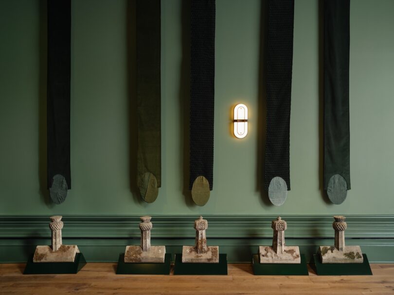 Six stone artifacts displayed on pedestals against a green wall with vertical dark fabric panels and a central wall light.