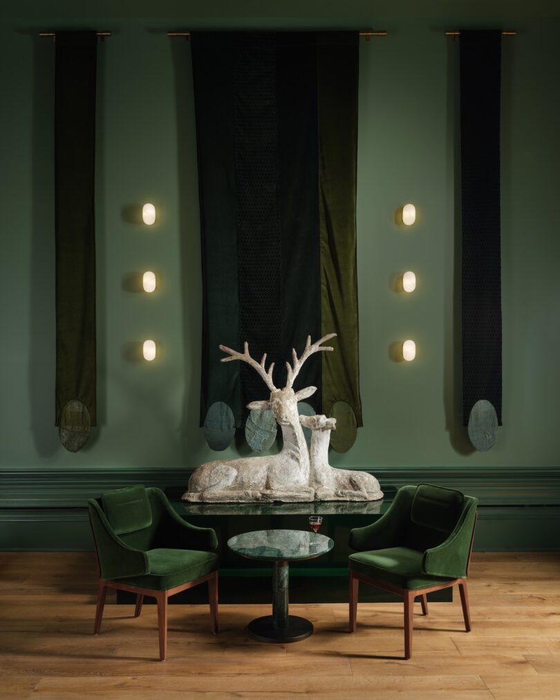 A green-themed room with a deer sculpture on a table, two green chairs, vertical fabric panels, and six small lights on the wall.