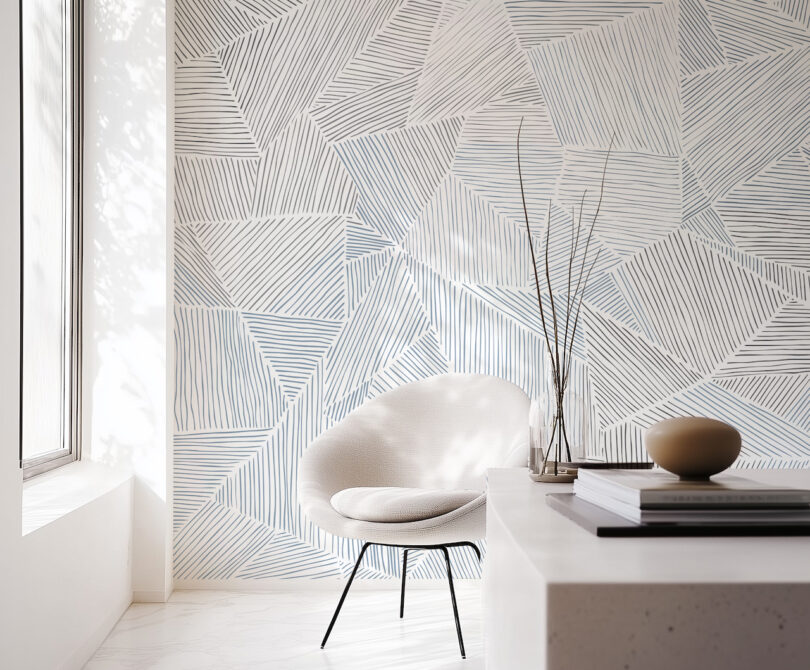 A white modern chair sits in a room adorned with Jill Malek's geometric-patterned wallpaper. A table with decorative items graces the foreground, while sunlight gently filters through a window