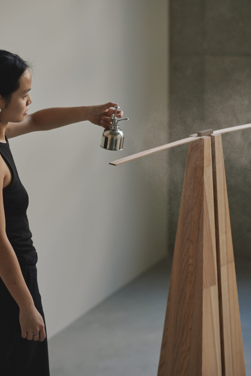 Person spraying a thin, flat wooden object with a metal spray bottle, next to a tall wooden stand in a minimalist room