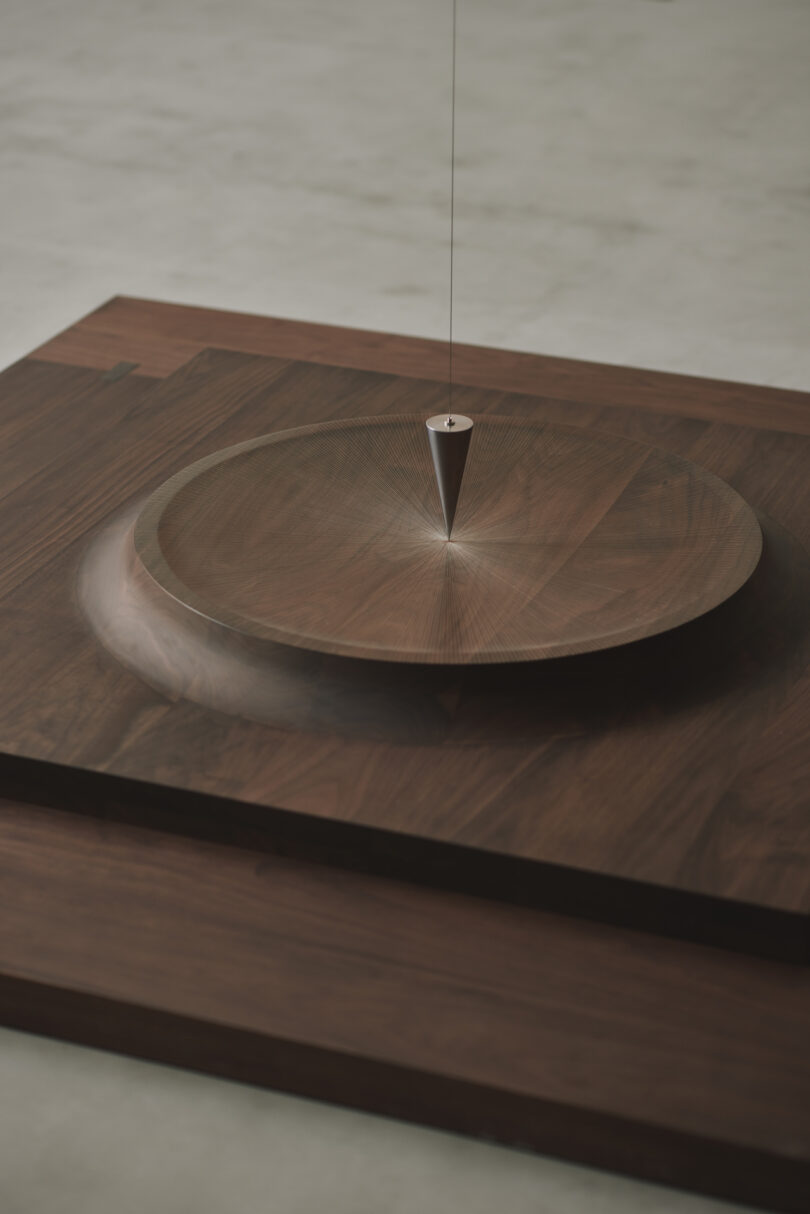 A metal pendulum hangs above a circular wooden platform on a square wooden base