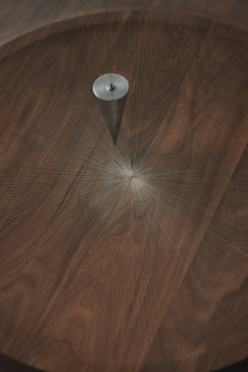 A metal pendulum hangs above a circular, intricately patterned wood surface