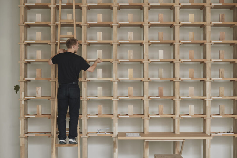 A person is standing on a ladder, arranging items on a large wooden shelving unit with multiple compartments