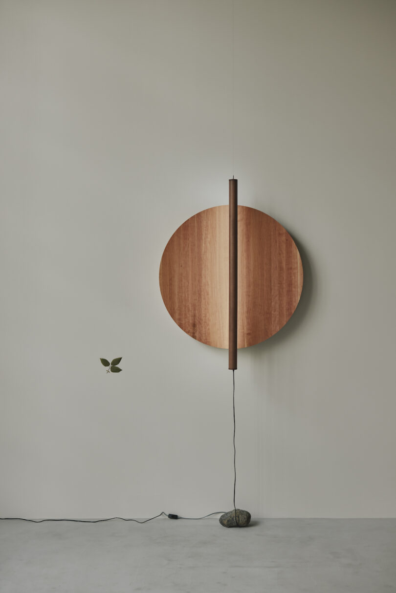 A wooden circular wall decoration with a vertical rod attached, supported by a stone base, is mounted on a plain light-colored wall. A small leafy branch is on the left side