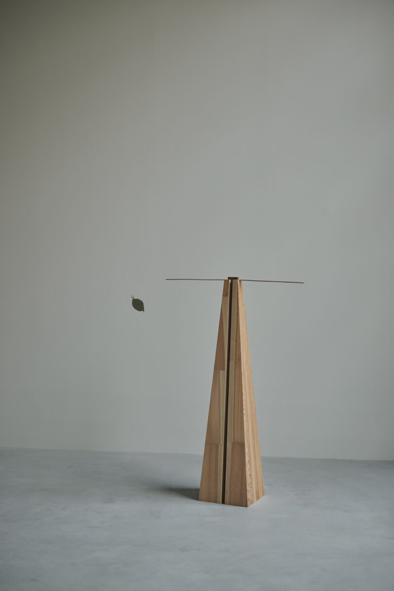 A minimalist wooden sculpture with a leaf suspended from a horizontal rod, set against a plain background