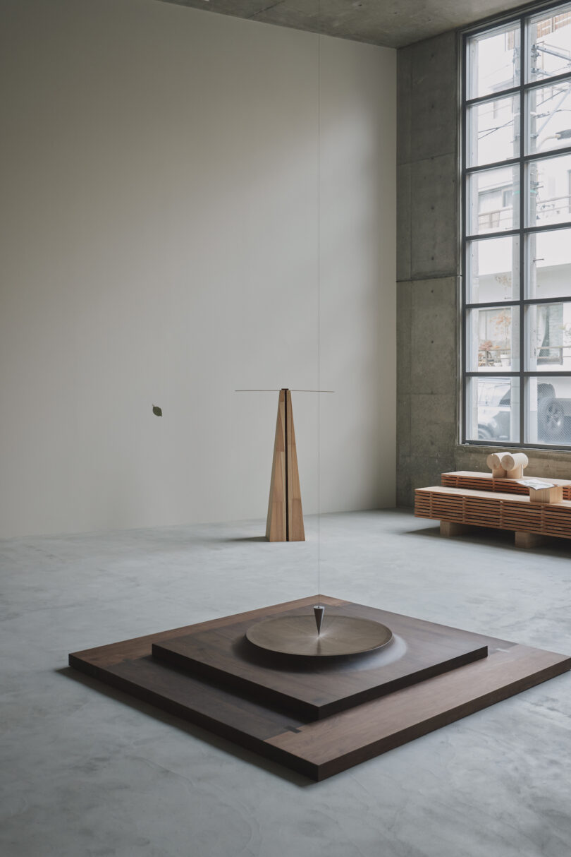 Minimalist room with a suspended pendulum over a circular wooden platform. Large window on the right and wooden benches with geometric objects in the background