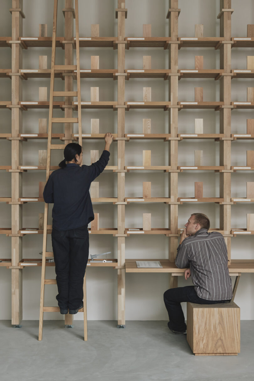 Two people organize a wooden shelf. One person stands on a ladder arranging items, while the other sits, writing notes