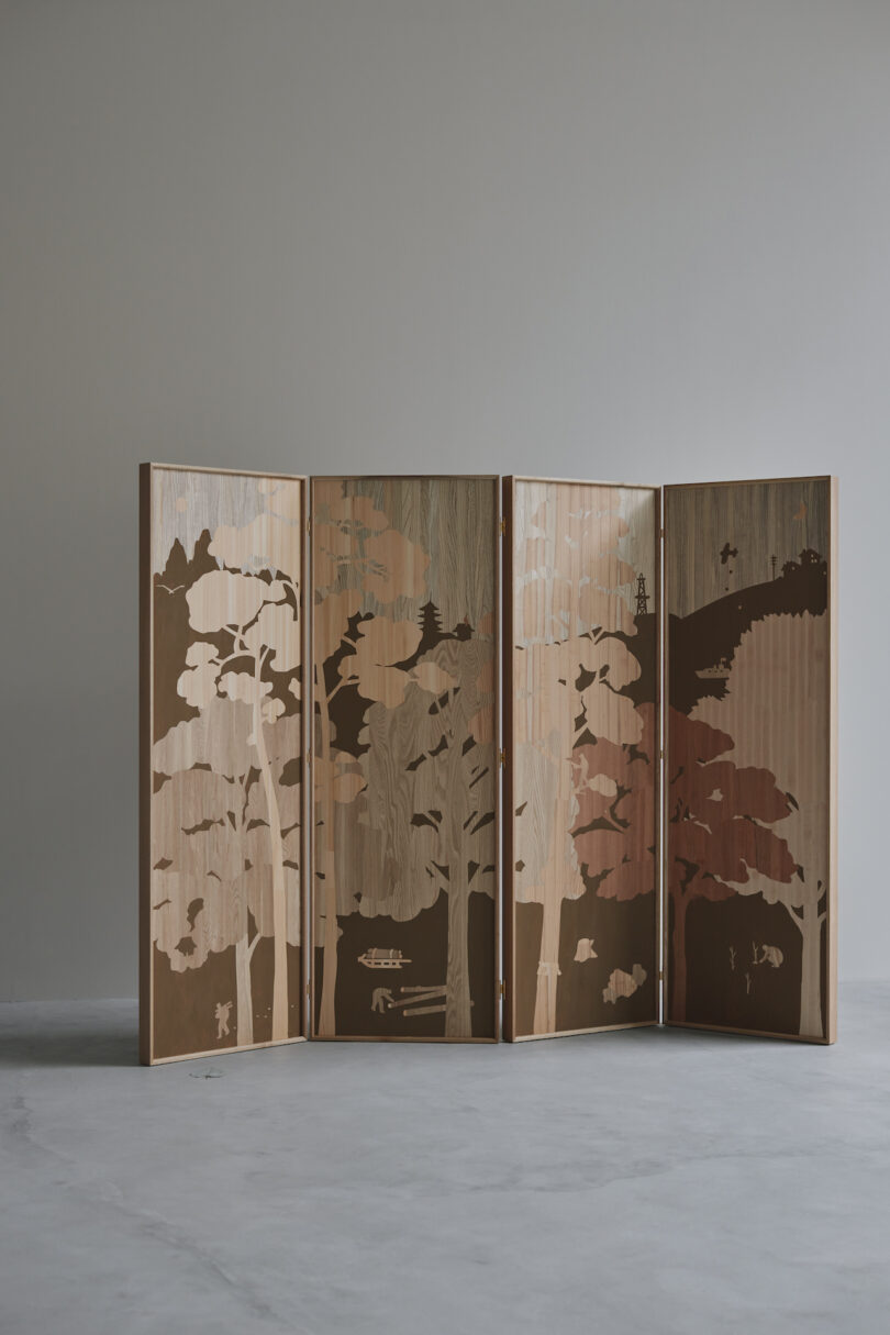 A decorative wooden folding screen with a nature-themed silhouette design stands against a plain gray wall on a concrete floor