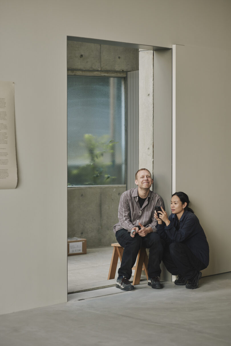 Two people sitting on a wooden bench, smiling and looking at a smartphone in a minimalist room with a large window