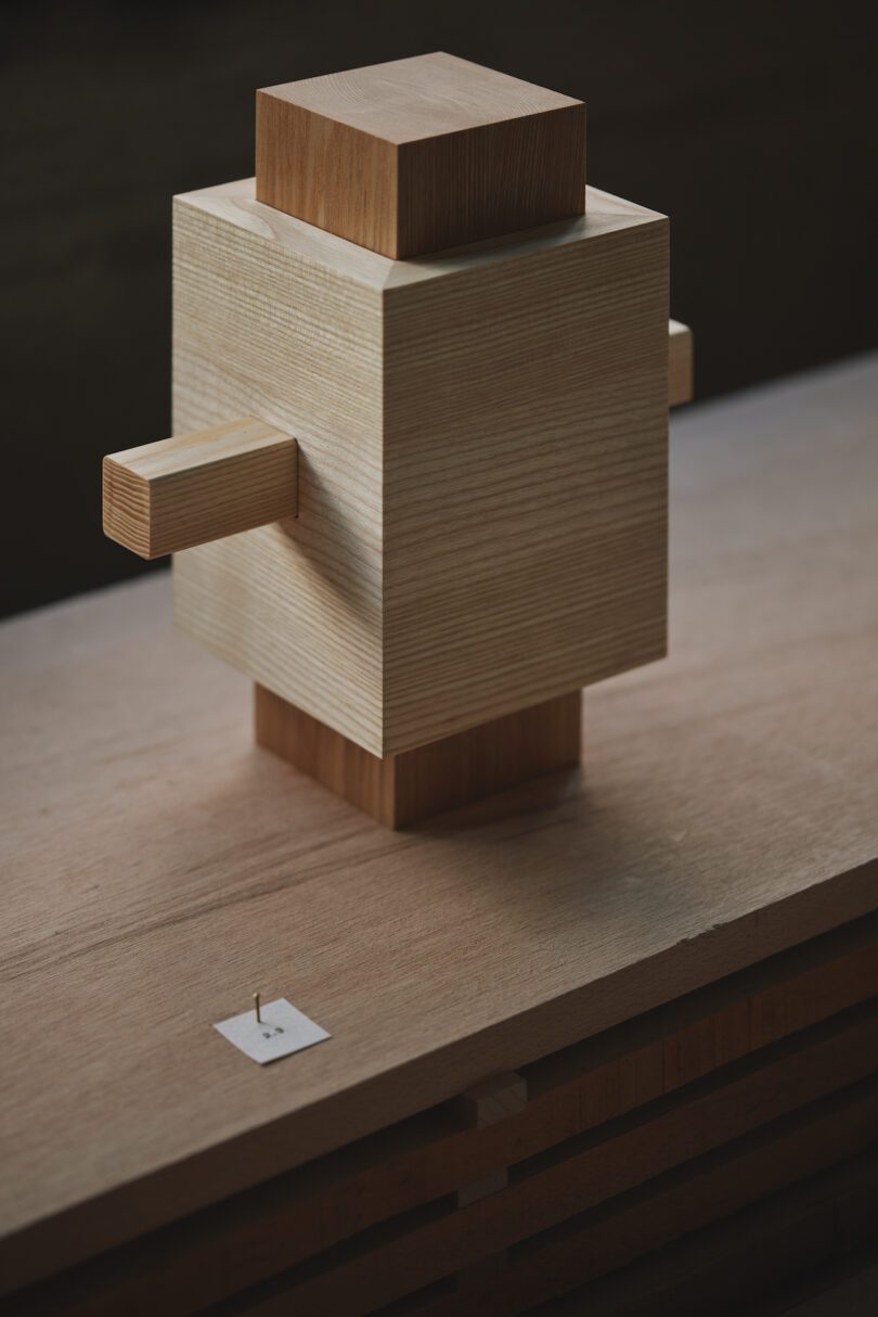 A wooden geometric sculpture with intersecting cubes and rectangular prisms sits on a wooden surface, lit from the side