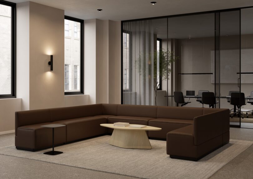 The modern office waiting area features a Keilhauer U-shaped brown sectional sofa, complemented by a wooden coffee table and a small side table. In the background, a glass-walled conference room enhances the sleek, neutral color scheme.