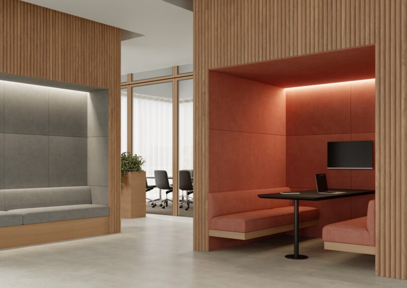 Modern office space with wooden accents, featuring a gray seating area, a red booth with a table and monitor, and a glimpse of a meeting room with chairs and a plant