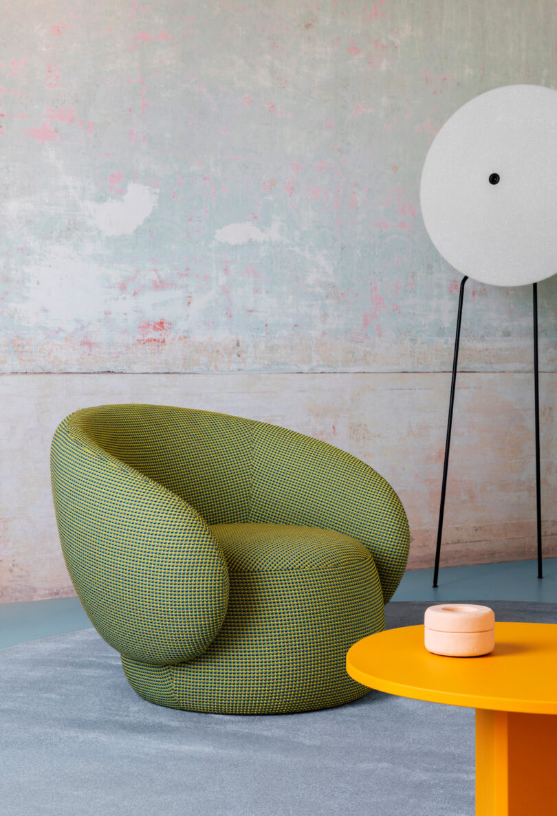 A green, modern curved chair sits next to a yellow table with a small round object on it. A tall, circular white lamp is in the background against a textured wall