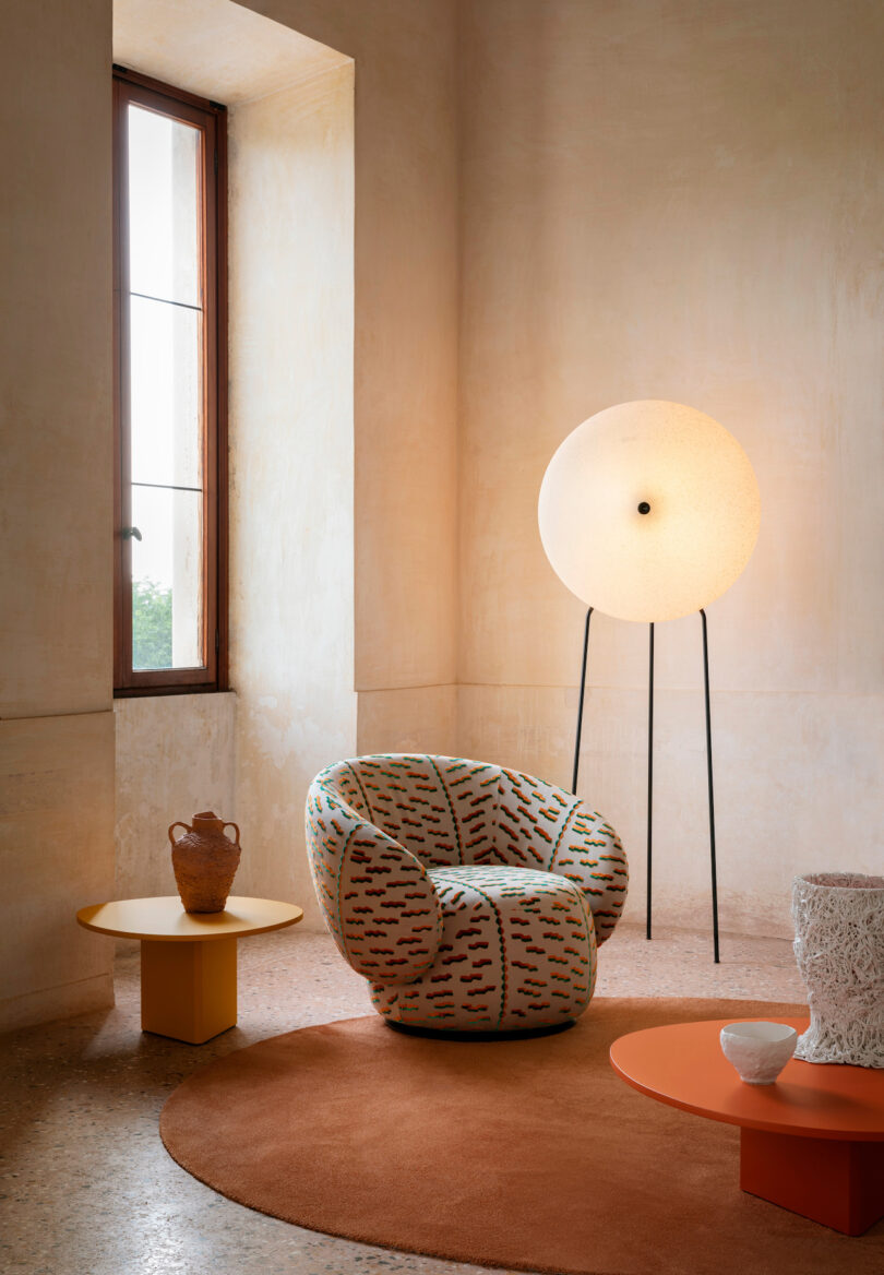 A cozy room with a patterned armchair, a tall spherical floor lamp, an orange rug, and two circular coffee tables with decorative items. A large window allows natural light in